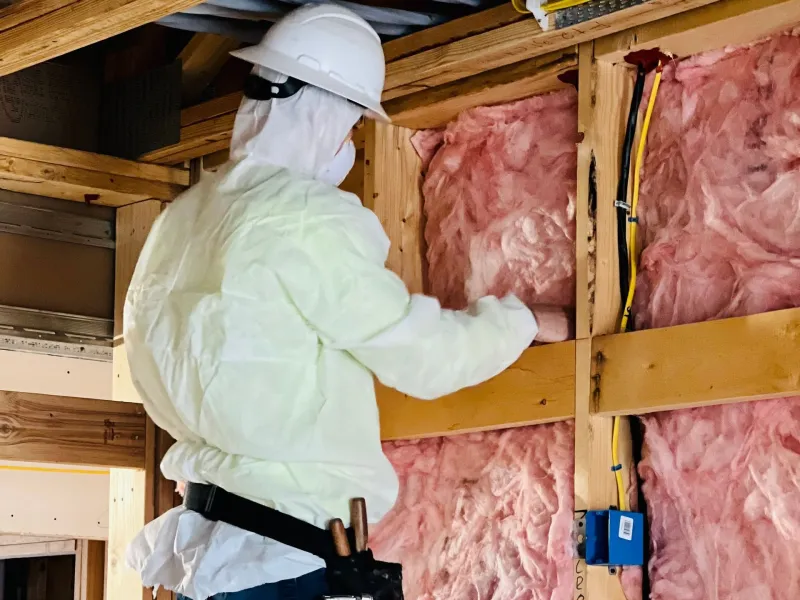 Installer in a hard hat fitting pink fiberglass batts between wall studs